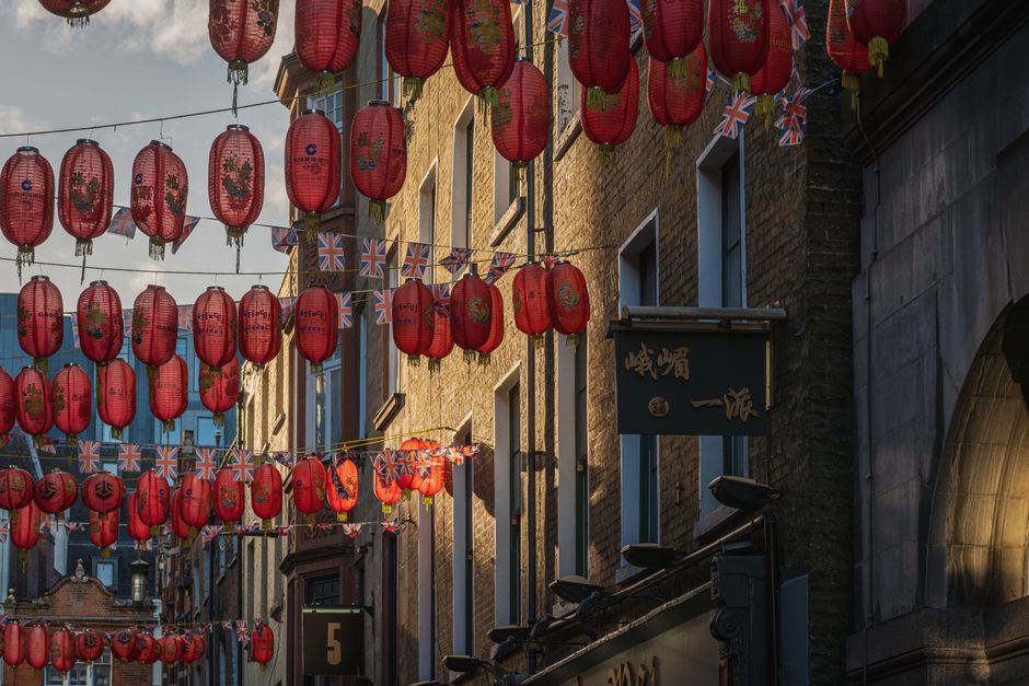 An outdoor street scene decorated with multiple rows of vibrant red Chinese lanterns and Union Jack flags hanging across the narrow alleyway. The lanterns, adorned with traditional gold floral pattern