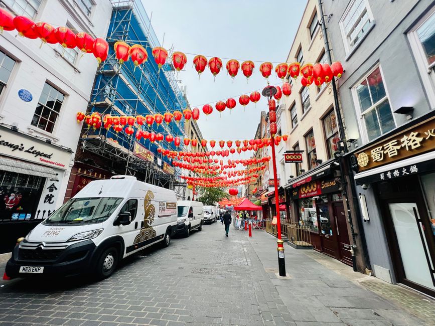 A street decorated with numerous strings of red Chinese lanterns hanging overhead, set against a backdrop of storefronts with Chinese signage and a building under scaffolding wrapped in blue safety ne